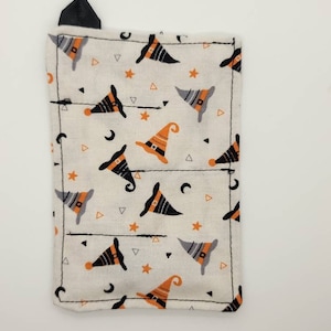 Könnte beinhalten: Eine kleine weiße Stofftasche mit einem schwarzen Reißverschluss. Die Tasche ist mit einem sich wiederholenden Muster aus orangefarbenen und schwarzen Hexenhüten, Sternen und Mondsicheln verziert.