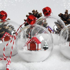 May include: Clear baubles with miniature winter scenes inside. Each bauble features a small house with a red roof, a tiny snowman, and a miniature Christmas tree, set in a snowy landscape. Red bells and striped string complete the festive look.