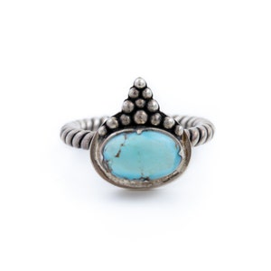 Op de afbeelding: Een zilveren ring met een turquoise steen en een getextureerde band. De ring heeft een uniek ontwerp met een puntige bovenkant en een parelmoerpatroon.