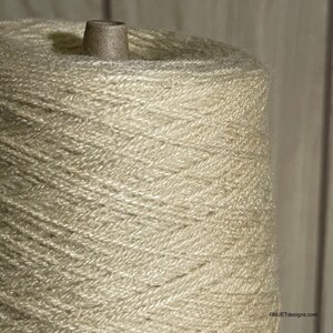 May include: Close-up of a large spool of off-white yarn. The yarn is tightly wound around a cardboard core, creating a conical shape. The yarn has a slightly textured appearance, suggesting a natural fiber. The background is a blurred, neutral tone.