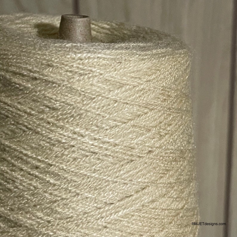 May include: Close-up of a large spool of off-white yarn. The yarn is tightly wound around a cardboard core, creating a conical shape. The yarn has a slightly textured appearance, suggesting a natural fiber. The background is a blurred, neutral tone.