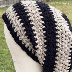 Peut inclure: Un bonnet crocheté fait à la main en noir et beige. Le chapeau présente des rayures verticales alternées de fil noir et beige. Le bonnet est présenté sur une tête de mannequin. Le texte "@RMJETDESIGNS" est visible dans le coin supérieur droit.