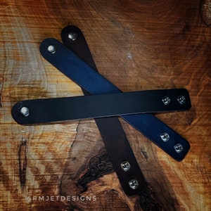 May include: Four leather wristbands in black, navy blue, and brown, each with silver snap closures. The bands are arranged on a wooden surface, showcasing the different colours and textures of the leather. The wristbands are a simple, yet stylish accessory.