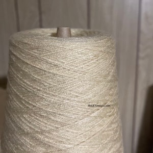 May include: A large spool of off-white yarn, tightly wound around a cardboard core. The yarn has a slightly textured appearance, with visible strands creating a spiral pattern. The image is well-lit, highlighting the yarn's color and texture. The text "RMJETdesigns.com" is visible.