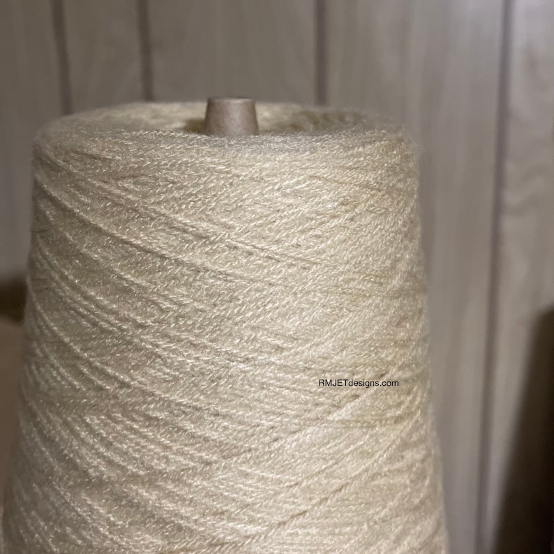 May include: A large spool of off-white yarn, tightly wound around a cardboard core. The yarn has a slightly textured appearance, with visible strands creating a spiral pattern. The image is well-lit, highlighting the yarn's color and texture. The text "RMJETdesigns.com" is visible.