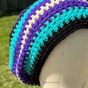 May include: A colorful crochet beret in shades of purple, teal, black, and cream. The handmade hat is displayed on a mannequin head. The beret features a striped pattern and is suitable for various occasions.