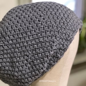 Peut inclure: Un bonnet en crochet gris. Le bonnet est fait avec un point texturé, créant un tissu dense et chaud. Le chapeau est présenté sur une tête de mannequin. Le texte "@RMJETdesigns" est visible dans le coin inférieur droit.