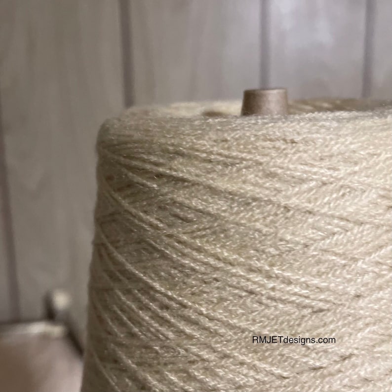 May include: Close-up of a large spool of beige yarn. The yarn is tightly wound around a cardboard core, creating a conical shape. The texture of the yarn is visible, suggesting a soft, natural fiber. The background is a light, neutral color.