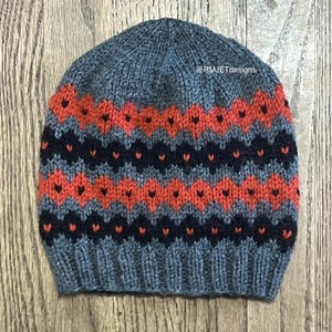 May include: A knitted beanie hat in blue, orange, and navy. The hat has a repeating pattern of orange and navy shapes against a blue background. The text "@RMJETdesigns" is visible on the hat.