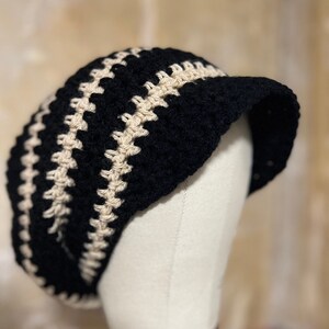 May include: A black and cream crochet beret with a visor. The hat has alternating bands of black and cream crochet stitches. The beret is displayed on a mannequin head.