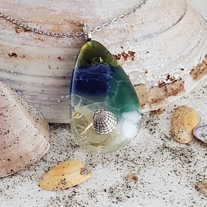 May include: A teardrop-shaped pendant necklace with a silver chain. The pendant is made of clear resin with layers of blue, green, and yellow, and a small silver seashell embedded in the center.