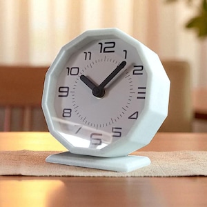 May include: A light blue, geometric-shaped analog alarm clock with a white face and black hands and numerals. The clock sits on a small, rectangular base. The clock is on a wooden table with a neutral-colored runner.