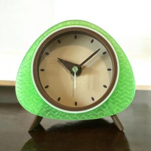 May include: A green and brown analog clock with a triangular base and three legs. The clock face is cream colored with black hour markers and black hands.