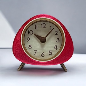 Puede incluir: Un reloj despertador rojo en forma de triángulo con detalles dorados y una esfera blanca. El reloj tiene agujas negras y números negros.