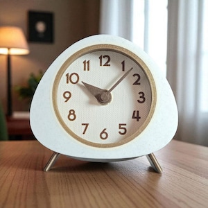 Puede incluir: Un reloj analógico blanco en forma de guijarro con un borde y patas doradas. La esfera del reloj muestra números y manecillas negros. El reloj mide aproximadamente 13 cm de ancho y 13 cm de alto.