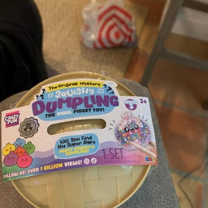 May include: A round, beige, woven-textured container holding a Squishy Dumpling fidget toy. The packaging features the text The Original Mystery Squishy Dumpling The Viral Fidget Toy, Series 3, 1 Set, and Will You Find the Super Rare Party Confetti Dumpling?