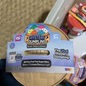 May include: A round, tan plastic container for a Mystery Dumpling toy, featuring a blue cardboard label with the text "The Original Viral Mystery Dumpling," "Silver Ribbon Edition," and "Will You Find The Super Rare Silver Ribbon Dumpling?"