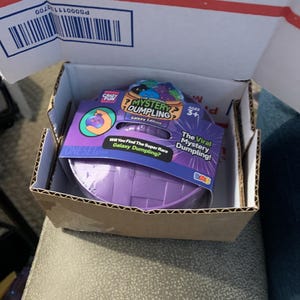 May include: A purple plastic basket container for The Original Mystery Dumpling Galaxy Edition toy set. The packaging features text including The Viral Mystery Dumpling, Will You Find The Super Rare Galaxy Dumpling, and Ages 3+ on a purple label.