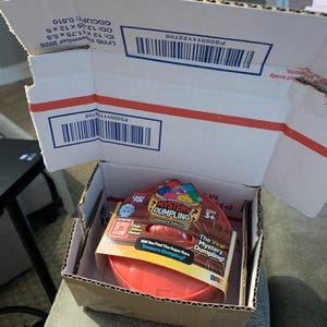 May include: A red plastic Mystery Dumpling Treasure Edition toy container inside a cardboard shipping box. The packaging features text including The Original Viral Mystery Dumpling, Treasure Edition, Ages 3+, and Will You Find The Super Rare Treasure Dumpling?