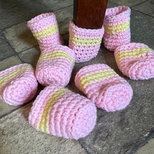 May include: A collection of seven handmade crochet furniture leg covers featuring a repeating pattern of soft pink and pale yellow horizontal stripes. Each cylindrical cover is crafted with a textured yarn stitch, designed to fit snugly around chair legs.