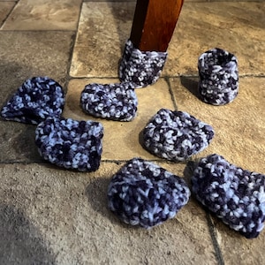 May include: A selection of handmade crochet chair leg protectors in blue, grey, and white. The protectors are in various shapes and sizes, with some fitted on a wooden chair leg. The yarn has a speckled appearance.