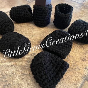 Può includere: Una collezione di protezioni per gambe di sedia nere, lavorate all'uncinetto. Le protezioni sono cilindriche e a forma di cappuccio, progettate per adattarsi alle gambe delle sedie. L'immagine include il testo "Little Gems Creations 1".