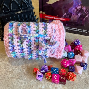 May include: A pink crocheted dice bag with a drawstring closure, filled with colorful dice. The dice are various shapes and sizes, with numbers printed on each side. The bag is sitting on a light brown surface.
