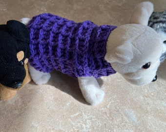 May include: A purple crocheted dog sweater on a white stuffed dog with a black stuffed dog in the foreground.