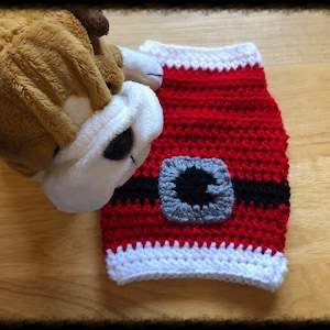 May include: A red and white crocheted dog sweater with a black belt and a grey buckle. The sweater is designed to look like a Santa Claus outfit.