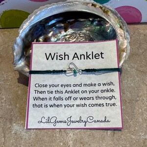 Può includere: Un braccialetto alla caviglia dei desideri con un cordino verde e un piccolo ciondolo argentato, presentato su una carta con istruzioni e la scritta "Wish Anklet". La carta è posta in una conchiglia iridescente.