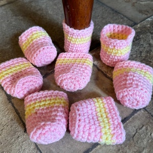 May include: A set of eight cylindrical crochet furniture leg covers featuring a light pink base color with a single horizontal yellow stripe encircling the middle. Each cover is crafted from yarn with a visible textured stitch pattern.
