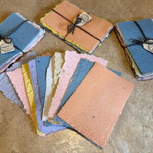May include: A collection of rectangular handmade paper sheets in various pastel colours including blue, peach, yellow, and lavender. The paper features rough, deckled edges and a textured surface. Three stacks are tied with black twine and white tags.