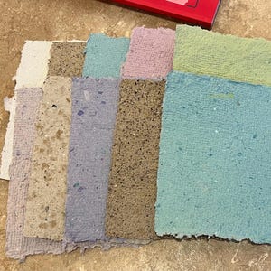 May include: A selection of handmade paper sheets in diverse colours, such as off-white, tan, light blue, pink, and green. The paper has a textured surface with visible flecks, indicating a natural, recycled composition. The sheets are arranged in a fan-like display.