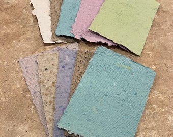 May include: A collection of rectangular handmade paper sheets with deckled edges, featuring a variety of colors including white, tan, light blue, lavender, and sage green. The paper has a textured, fibrous surface with visible flecks.