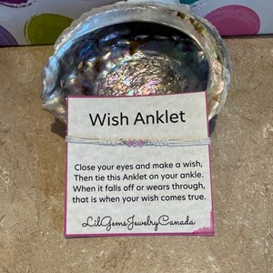 Puede incluir: Un brazalete de tobillo de cuerda blanca con cuentas rosas y una tarjeta que dice "Wish Anklet" y "Cierra los ojos y pide un deseo, Luego ata este brazalete de tobillo en tu tobillo. Cuando se caiga o se desgaste, ese es el momento en que tu deseo se hará realidad. Lil Gems Jewelry Canada".