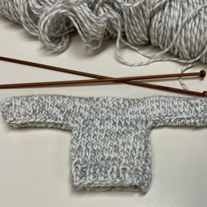 Puede incluir: Un suéter de punto gris y blanco con mangas largas, colocado plano sobre una superficie blanca. También se muestran dos agujas de tejer de madera.