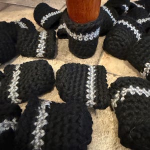 May include: A collection of black crochet chair leg covers featuring a single horizontal silver stripe. Each cover is constructed from thick yarn with a textured knit pattern, designed to fit snugly around the base of a wooden furniture leg.