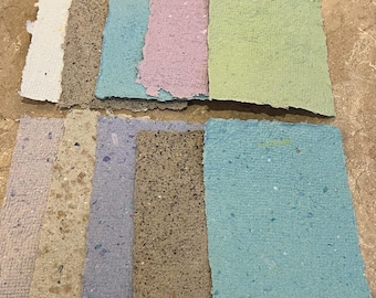 May include: A collection of handmade paper sheets in various colors, including white, brown, blue, pink, and green. The paper has a textured surface and irregular edges, suggesting a natural, artisanal quality. Ideal for crafts and stationery.