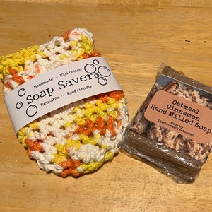 May include: A crocheted soap saver in white, orange, and yellow, with a label that reads "Soap Saver" and "Handmade... 100% Cotton Reusable... Ecofriendly". Next to it is a bar of oatmeal cinnamon hand-milled soap wrapped in plastic.