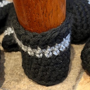 May include: A black crocheted furniture leg cover featuring a single horizontal stripe of light gray yarn. The cylindrical sleeve is fitted snugly around a wooden furniture leg, showcasing a textured, hand-stitched pattern throughout the design.