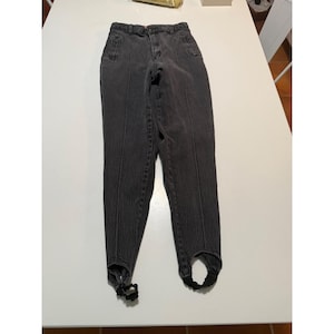 May include: High-waisted stirrup pants in a faded charcoal gray denim fabric. These pants feature a structured waistband with a button and zipper closure, vertical seam detailing running down the front of each leg, and black elastic stirrups at the cuffs.