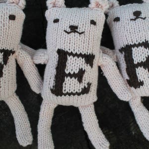 May include: Three handmade, light pink knitted bear dolls with dark brown embroidered features. Each bear has a letter on its chest: "E", "R", and "B". The bears have small ears, arms, and legs. The background is a dark, textured wooden surface.