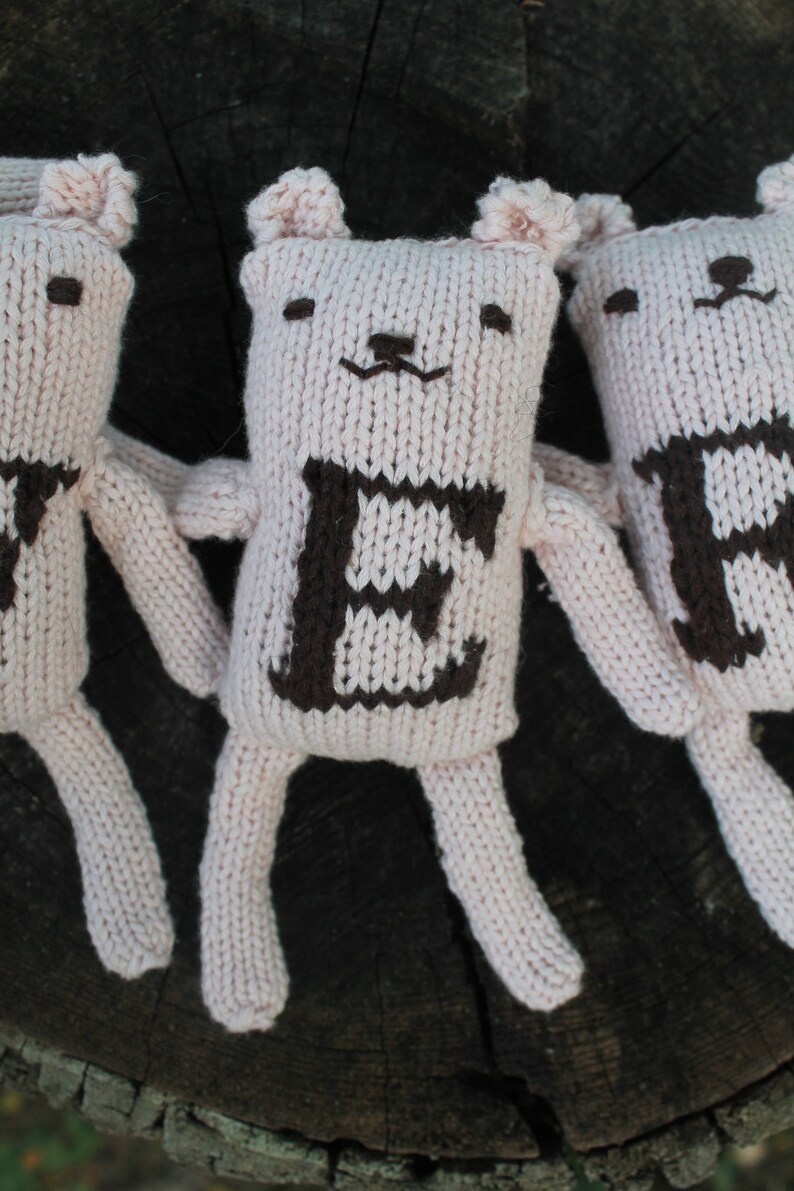May include: Three handmade, light pink knitted bear dolls with dark brown embroidered features. Each bear has a letter on its chest: "E", "R", and "B". The bears have small ears, arms, and legs. The background is a dark, textured wooden surface.