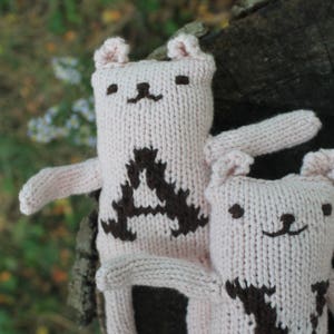 May include: Two light pink knitted bear dolls with brown embroidered details. One bear has a large brown "A" on its chest. The other bear has a brown "V" on its chest. The bears have small ears, arms, and smiling faces.