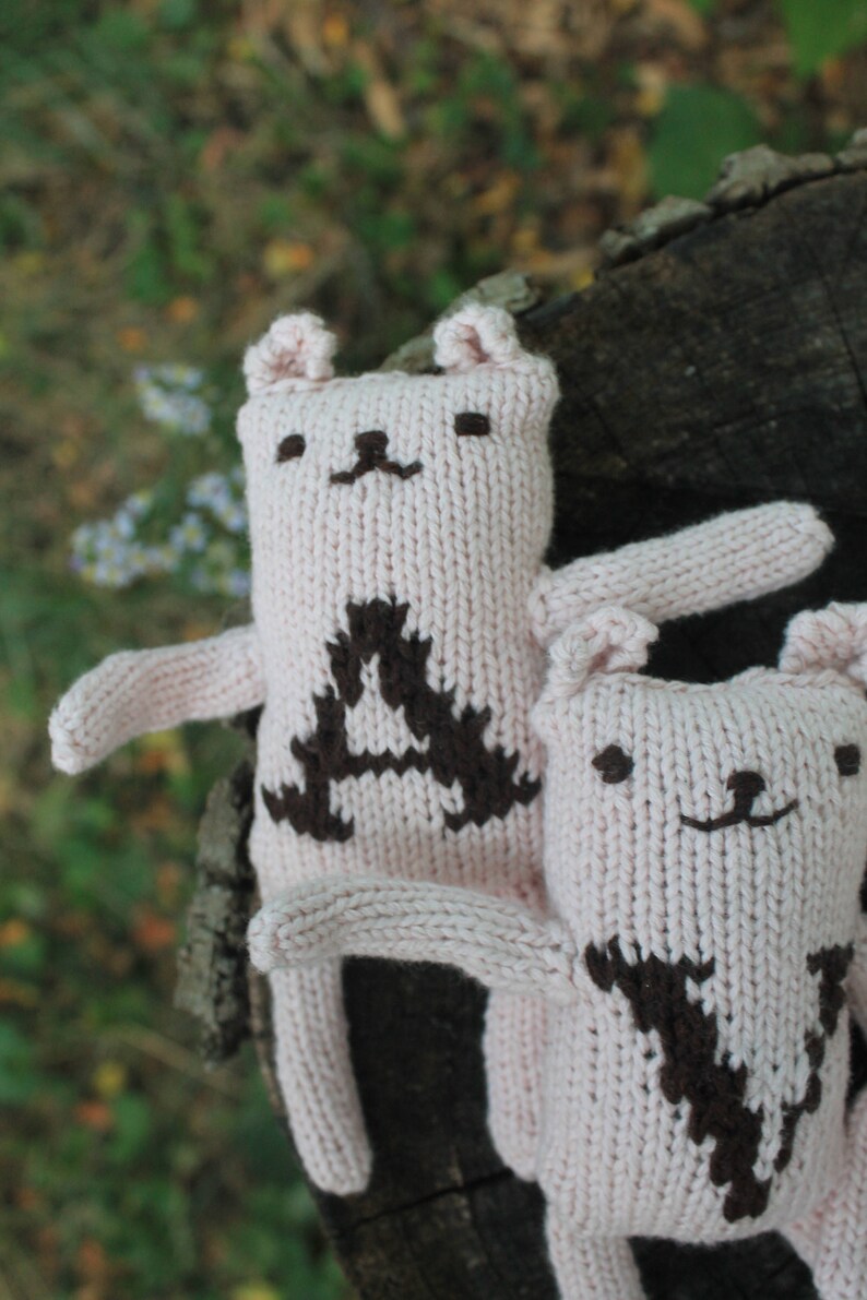 May include: Two light pink knitted bear dolls with brown embroidered details. One bear has a large brown "A" on its chest. The other bear has a brown "V" on its chest. The bears have small ears, arms, and smiling faces.
