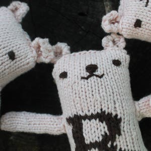 May include: Close-up of several handmade, knitted bear dolls. The bears are primarily light pink with brown facial features and a brown pattern on the chest. The dolls have small, rounded ears and long arms.