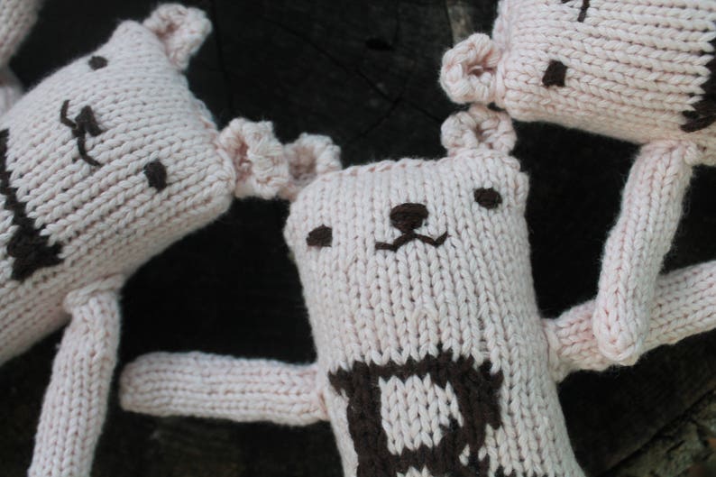 May include: Close-up of several handmade, knitted bear dolls. The bears are primarily light pink with brown facial features and a brown pattern on the chest. The dolls have small, rounded ears and long arms.