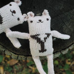 May include: Several handmade, light pink knitted bear dolls with brown details. The bears have small ears, arms, and legs. One bear is in focus, with a brown Y-shaped design on its chest. The dolls are on a dark wood surface.