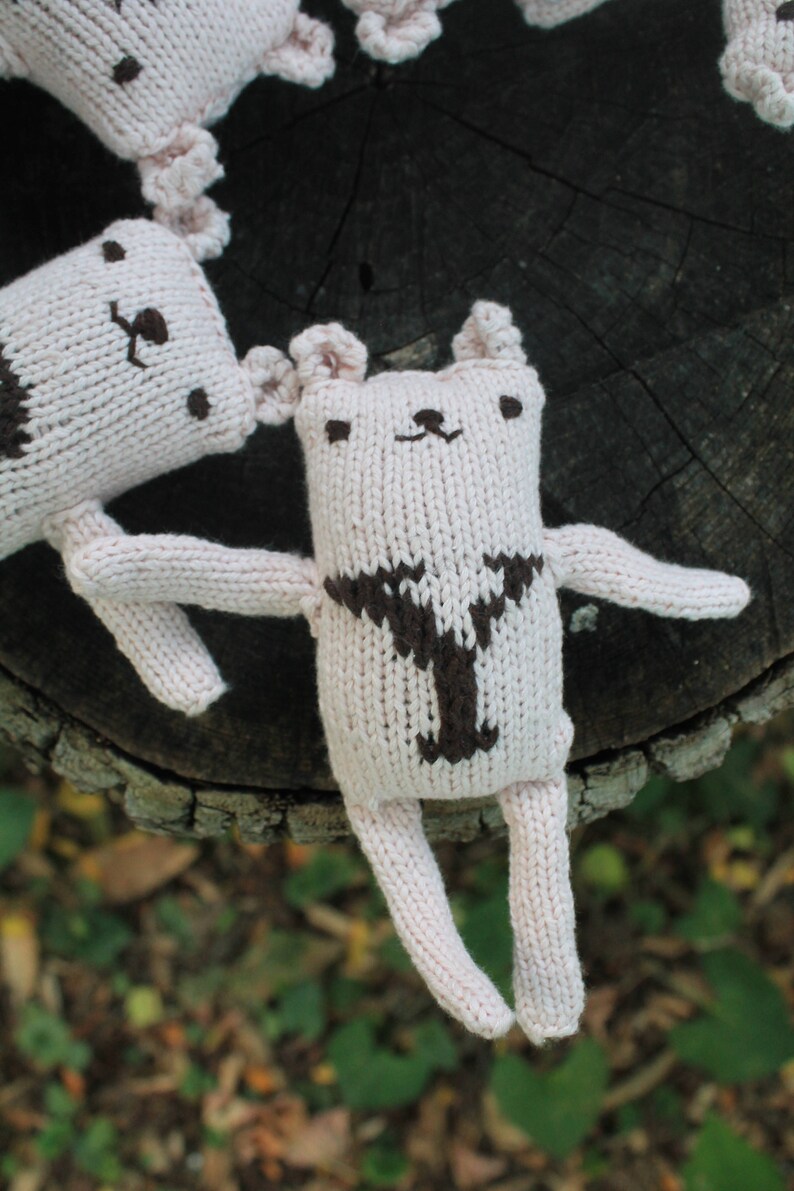 May include: Several handmade, light pink knitted bear dolls with brown details. The bears have small ears, arms, and legs. One bear is in focus, with a brown Y-shaped design on its chest. The dolls are on a dark wood surface.