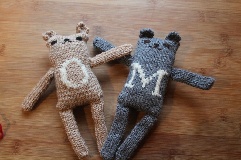May include: Two handmade knitted bear dolls. One is tan with a white "O" on its chest, the other is gray with a white "M". Both have small ears, arms, and legs. They are lying on a wooden surface.
